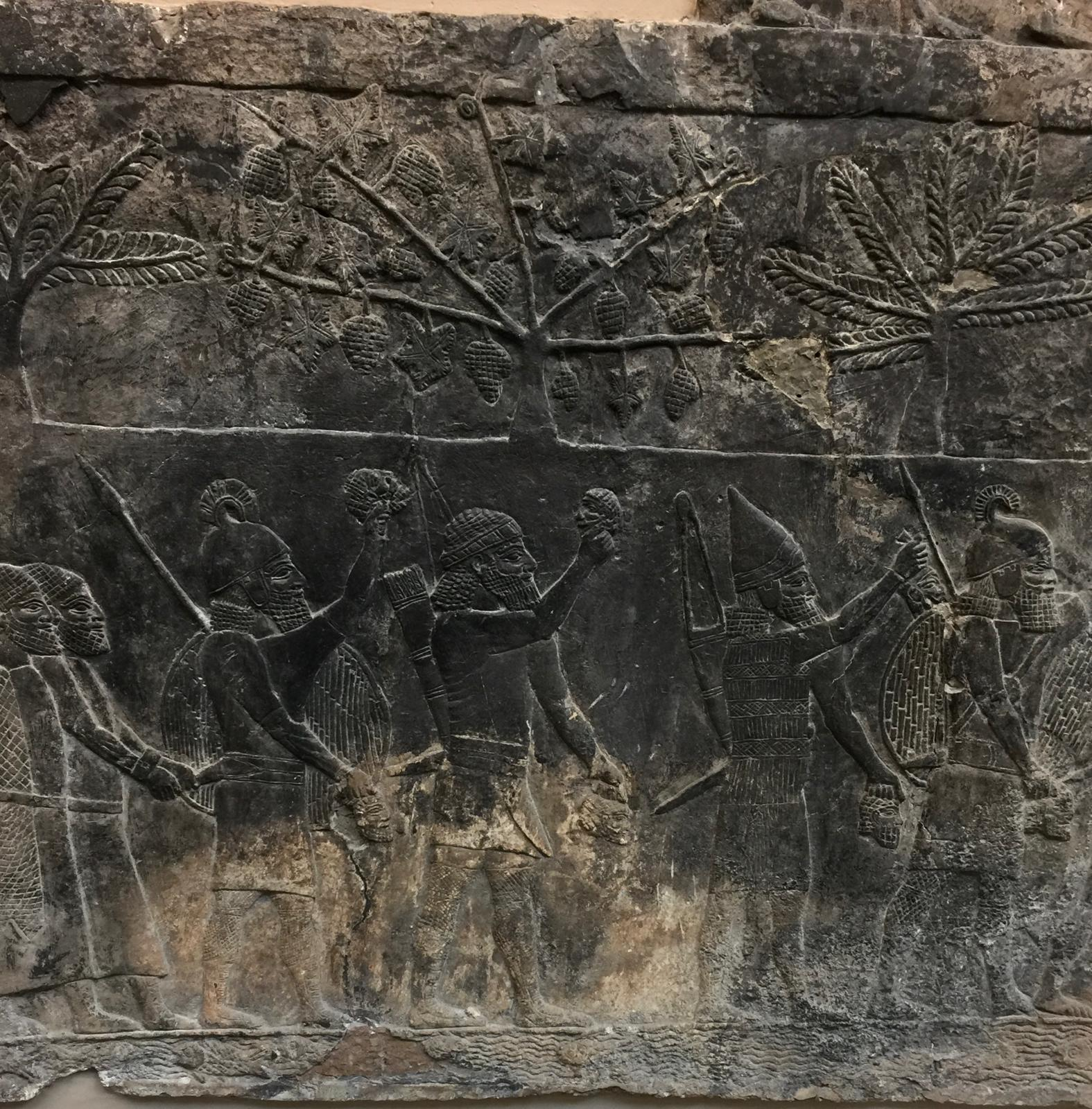 A stone relief from Nineveh showing Assyrian soldiers carrying severed heads beneath fruit trees and palms.